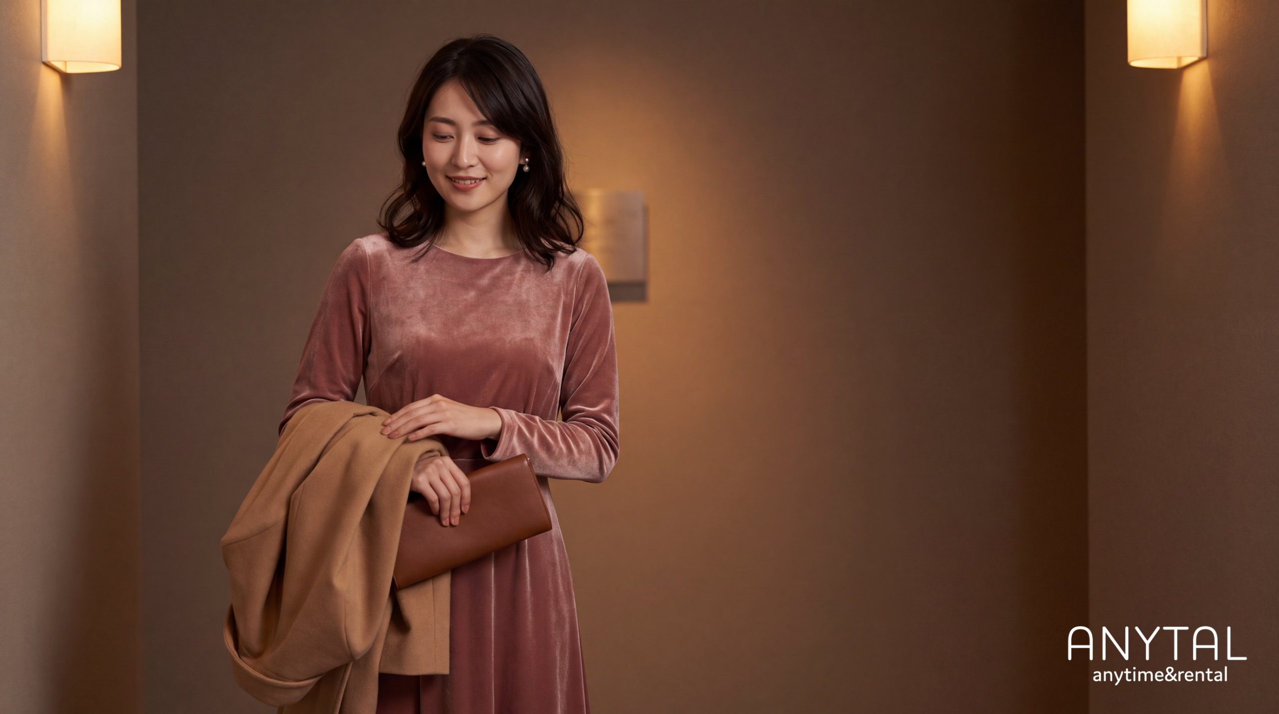 くすみピンクの長袖ドレスを着て、ブラウンのコートとバッグを持つ女性